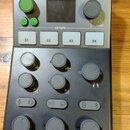 Černý mixážní stolek s horní stříbrnou částí a displejem. V horní části je nápis M-CASTER STUDIO s logem. Zařízení obsahuje čtyři bílá tlačítka označ…