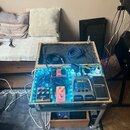 Otevřený dřevěný kufr s vnitřním polstrováním, který slouží jako pedalboard. Uvnitř jsou rozmístěny různé kytarové efekty, které jsou podsvícené modr…
