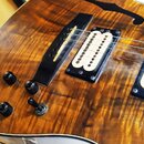 Detail těla elektrické kytary s výraznou dřevěnou masivní strukturou v hnědých a žlutých odstínech. Nástroj má dva humbuckery s bílými plastovými kry…