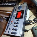 Stříbrný sampler/sekvenční stroj s nápisem MACHINEDRUM a logem Elektron. Zařízení má displej, několik řad tlačítek a šedé otočné knoflíky. Na pravém…