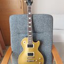 Zlatá elektrická kytara typu Les Paul s dvěma humbuckery a čtyřmi ovládacími knoflíky. Na hlavě je bílý nápis Epiphone a zlatý nápis Les Paul. Kytara…