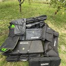 Hromada černých textilních obalů a přepravních braš různých velikostí a tvarů položená na trávě. Na několika kusech je vidět bílý nápis ROCKBAG a na…