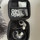 Otevřený černý přepravní pouzdro s vnitřní síťovkou, ve které jsou uloženysilikonové nástavky. V pouzdře se nacházejí in-ear monitory s průhledným tě…