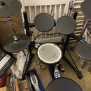 Elektronická bicí souprava značky Roland s černými pady a činely. Na module je nápis Roland TD-5 V-Drums. Centrální pad má bílý povrch s logem Roland…