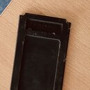 Černý obdélníkový plastový nebo kovový kousek příslušenství s vyhlubní uprostřed. V horní části je vrytý nápis 156055. Povrch vykazuje známky opotřeb…