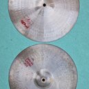 Dvě kovové činely v stříbrné barvě s viditelným broušením a otisky kladiva. Horní činel nese nápisy HEAVY HI-HAT BOTTOM 14" a logo Paiste 2002. Dolní…