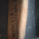 Detailní záběr na část černého povrchu s přilepeným nebo vtištěným světle hnědým štítkem. Na štítku je svisle napsáno MODEL L75 a pod tím kód 378-040…