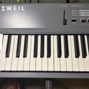 Šedý digitální klavír s bílými a černými klávesami. Na levé straně je nápis KURZWEIL a dvě černé ovládací kolečka. V horní pravé části jsou různé pře…
