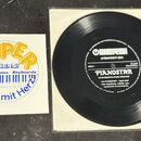 Fotografie zobrazuje černý vinylový disk v bílém obalu a doprovodný čtverečkový papír s reklamním logem. Na bílém papíře je žlutý nápis SUPER, logo W…