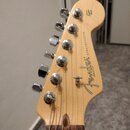 Fotografie zachycuje hlavu elektrokitary z přírodního světlého dřeva. Na povrchu je čitelné logo Fender, nápis STRATOCASTER a drobný text MADE IN U.S…