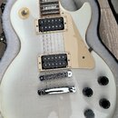 Bílá elektrická kytara s tvarem singlecut v otevřeném kufru s bílou výplní. Nástroj má dva humbuckery, čtyři ovládací knoflíky a světle béžový plecht…