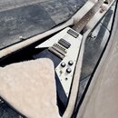 Černá elektrická kytara s trojúhelníkovým tělem a bílým pickguardem v otevřeném tvrdém kufru s bílou výstýlkou. Nástroj má dva humbuckery, Tune-o-mat…