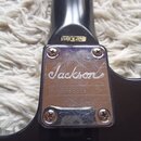 Záhlaví černé kytary s kovovým krytem krkovým spojem. Na hřidně je nápis MADE IN JAPAN a na kovovém krytu logo Jackson oraz sériové číslo 9832181. Po…