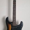 Elektrická kytara typu Stratocaster s tmavě hnědým přechodem do žluté barvy (sunburst) a černým pickguardem. Na hlavě hmatníku je vidět nápis "Squier…