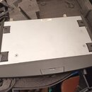 Fotografie zachycuje spodní stranu šedého elektronického zařízení v přenosném kufru s pěnovou výplní. Na spodním kovovém panelu jsou čtyři gumové nož…