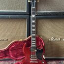 Červená elektrická kytara s černým pickguardem a bílými vložkami na hrazdici. Na hlavě je viditelné logo Gibson. Kytara je umístěna v otevřeném červe…