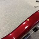 Detailní záběr na červený lakovaný povrch hudebního nástroje. Na hraně je vidět výrazný odlakovaný škrábanec a menší rýha v laku. V pravé části je vi…