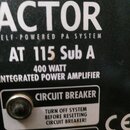 Detailní záběr na zadní panel černého aktivního subwooferu. Je zde uveden nápis ACTOR, ELF-POWERED PA SYSTEM a model AT 115 Sub A. Dále je vidět text…