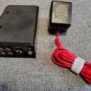 Černý kovový box s několika konektory na zadní straně. Jsou zde vidět dva zlaté konektory s nápisy „Out Ph1“ a „Out Ph2“, jeden černý konektor „Out X…