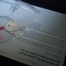Na bílém papíře s textem v němčině a logem „MUSIK MEYER“ leží malý kovový díl v průhledném plastovém obalu. Na papíře je vidět kulaté razítko s nápis…