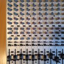 Fotografie znázorňuje míchací pult stříbrné barvy s mnoha ovládacími prvky. V horní části je nápis BEHRINGER a text ULTRA-LOW NOISE DESIGN. Na panelu…
