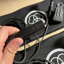 Černý mikrofon s dlouhým tenkým kabelem, který drží ruka. Na těle mikrofonu je vyražený nápis "AKG". V pozadí jsou dvě černé textilní pouzdra s bílým…