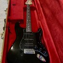 Černá elektrická kytara s trestým tělem a černým pickguardem, položená v červeně vystlaném kufr. Na hlavě hředu je viditelné logo Fender. Kytara má t…
