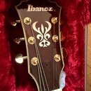 Detail hlavy kytary v tmavě hnědé barvě s bílým lemováním, umístěné v červeném plyšovém pouzdře. Na horní části je bílý nápis Ibanez a uprostřed je u…