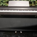 Digitální piano v tmavém dřevěném provedení s bílými a černými klávesami. Na notovém stojanu je nápis CELVIANO. Nástroj stojí na odpovídajícím černém…