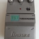 Stříbrný kytarový efekt pedál s nápisy TS7 TUBESCREAMER a logem Ibanez. Na horní části jsou tři ovládací knoflíky s popisky DRIVE, TONE a LEVEL a pře…