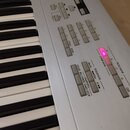 Detailní pohled na ovládací panel bílého klávesového nástroje. Jsou vidět různé sady tlačítek a nápisy jako COMB BANK, PROG BANK, SEQUENCER, SAMPLING…