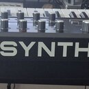 Černý synthesizér s bílými bočními panely. Na přední straně je oranžové logo ASM a bílý nápis HYDRASYNTH. Horní panel obsahuje klávesnici, početné ot…