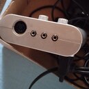 Stříbrné elektronické zařízení s kovovým povrchem, které obsahuje jeden velký kruhový konektor a tři menší jackové vstupy. Na horní straně jsou vidět…