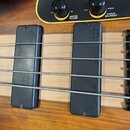 Detail basové kytary s dvěma černými snímači. Na snímačích je viditelný nápis "bactoline mb1". Ovládací panel je žluté barvy s nápisy "BASS BOOST/CUT…