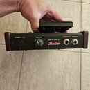 Zadávací panel černého elektronického zařízení s hnědým okrajem. Jsou zde nápisy POWER --( )+, VOLUME, STEREO/R a MONO/L. Uprostřed je bílý nápis MAD…