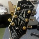 Hlava elektrické kytary v černé barvě s bílým lemováním. V horní části je nápis Epiphone a v dolní části nápisy Les Paul CUSTOM. Struny jsou upevněny…