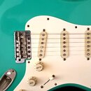 Detail elektrické kytary s tyrkysovým bodies a bílým pickguardem. Nástroj má tři bílé snímače typu single-coil, chromovaný kobylník a hnědý hmatník.…