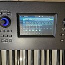 Detailní pohled na ovládací panel a klávesnici elektronického nástroje v šedé barvě. Uprostřed je umístěn displej s nápisem Performance a vybraným pr…