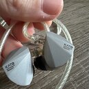 Dvě šedé sluchátka s hranatým, geometrickým tvarem a průhledným opleteným kabelem. Na obou sluchátkách je černý nápis KATO® MOONDROP PRESENTS. Povrch…