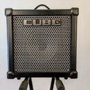 Černý kytarový kombozesilovač s kovovou mřížkou reproduktoru. V horní části mřížky je umístěn nápis CUBE. Nástroj má v rozích ochranné plastové rohy…