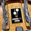Detail na korpus bubnu z dřeva s přirozenou Maserací. Uprostřed je černý štítek s bílým logem dvou kruhů, nápisem SONORLITE a textem No. 523 MADE IN…