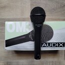Černý dynamický mikrofon s mřížkou, na těle kterého je bílý nápis AUDIX OM3. Mikrofon leží na originální krabici, na které jsou vidět nápisy OM3 a AU…