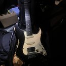 Černá elektrická kytara s bílým pickguardem a světle hnědým hmatníkem. Na hlavě nástroje je viditelné logo Fender. Tělo kytary má v dolní části výraz…