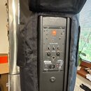 Černý aktivní reproduktor v černém transportním obalu. Na zadním panelu je oranžové logo JBL a nápisy PROFESSIONAL, EON 615 a MASTER VOLUME. Jsou zde…