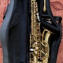 Zlatý saxofon v černém měkkém pouzdře. Nástroj je vybaven bílými polštářky a kovovými klapkami, na těle nejsou viditelné žádné značky ani nápisy. Ved…