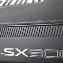 Detailní záběr na černý klavír s mřížkou reproduktoru a žebrovaným prvkem. Na spodní části je bílý nápis PSR-SX900. Povrch vykazuje drobné znečištění…
