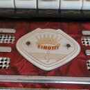 Detail červeného akordeonu s bílým plastovým štítkem ve tvaru štítu. Na štítku je zobrazen nápis FIROTTI, nad ním stylizovaná koruna a pod nápisem sy…
