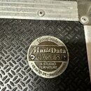 Detail kovového štítku na černém povrchu s protiskluzovým vzorem. Štítek nese nápisy „Custom-made“, „MusicData CASES“ a „& STUDIO FURNITURE“. Dole je…