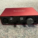 Červená audio rozhraní s nápisem Focusrite na horní straně a Scarlett Solo v levém dolním rohu. Zařízení má dva otočné knofíky, jeden velký ovládací…