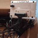 Fotografie zachycuje zadní stranu stříbrného elektronického zařízení značky Roland, které leží v kartonové krabici s nápisem BATA GOES GREEN. Na zadn…